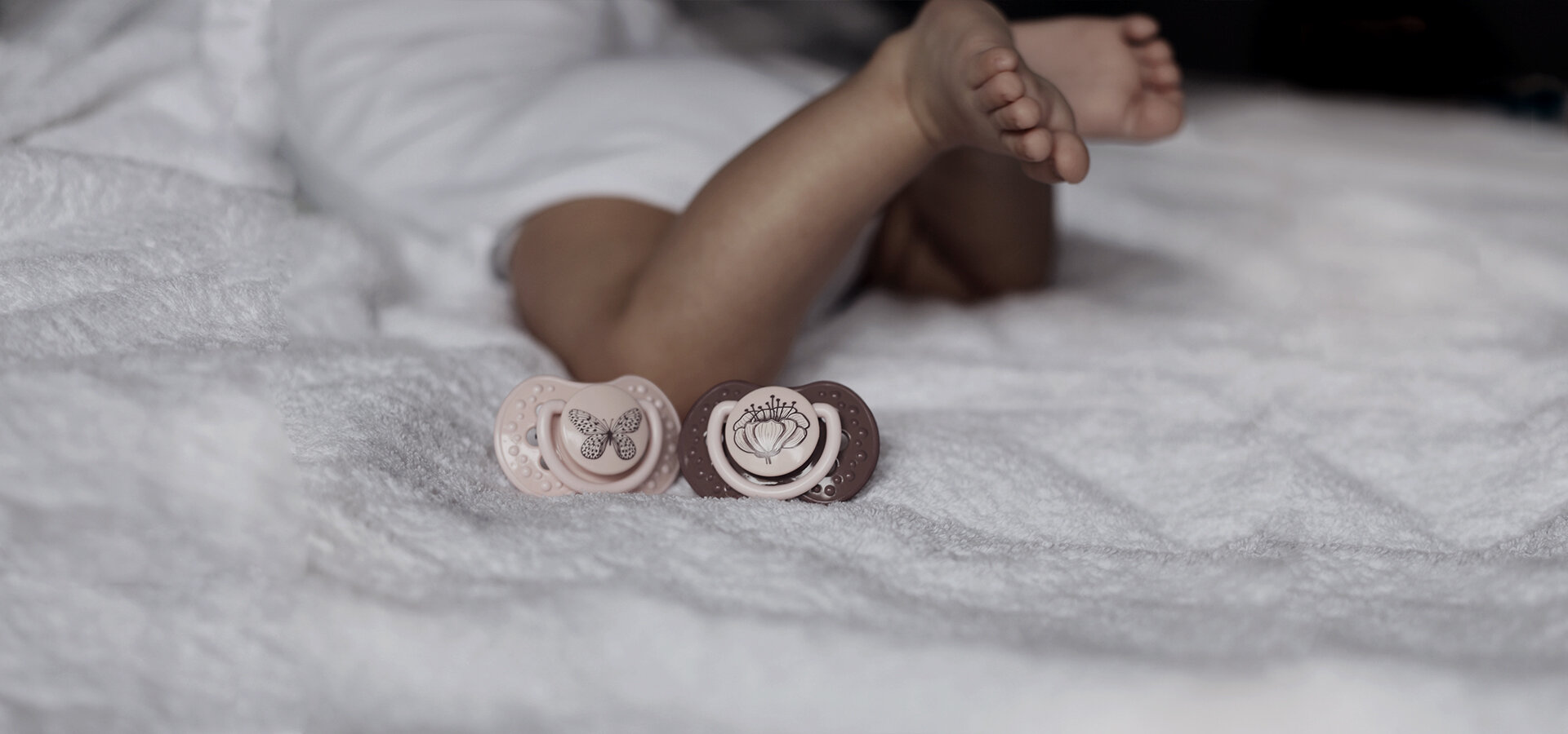 Ребенок лежит на кровати с двумя сосками-пустышка lovi из коллекции Botanic